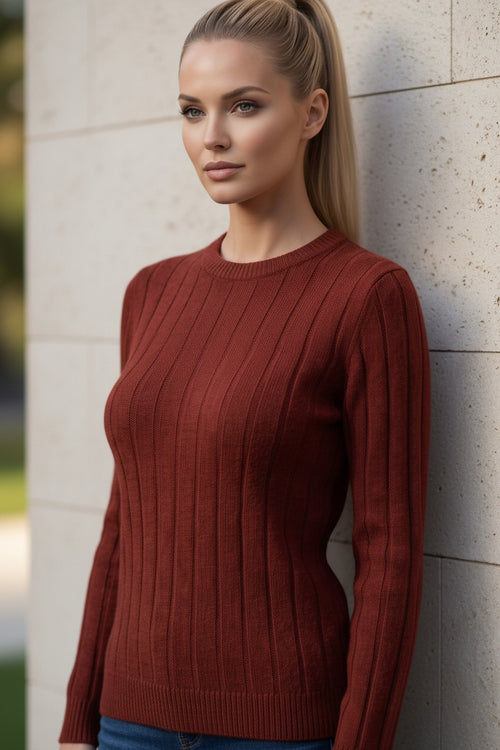 A woman with blonde hair in a ponytail wearing a rust-colored ribbed knit sweater, leaning against a gray brick wall in warm sunlight.