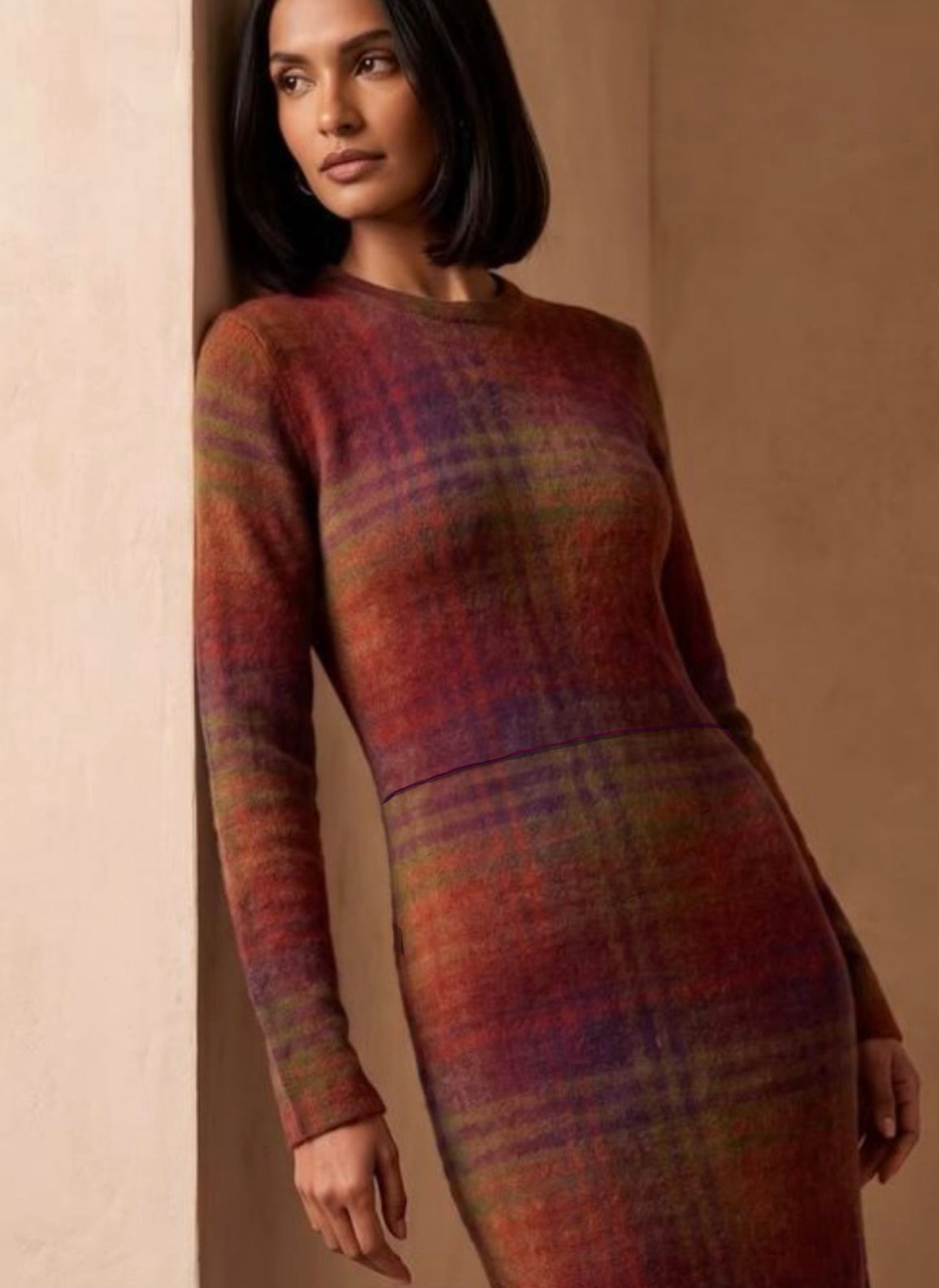 A woman wearing a fitted long-sleeve knit dress with a muted plaid pattern in red, purple, green, and orange tones, posed against a warm beige wall.