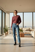 A woman modeling a maroon long-sleeve knit sweater and blue jeans with black ankle boots, standing against a light gray studio background.