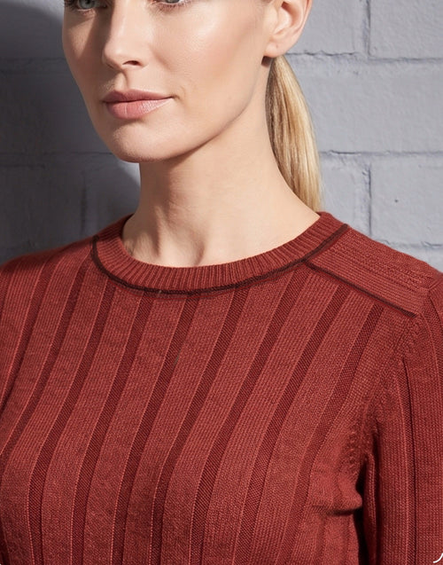 A close-up of a woman wearing a rust-red ribbed knit sweater against a gray brick wall.