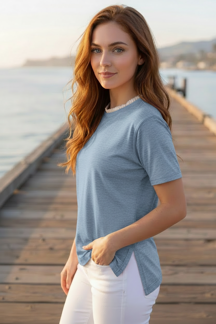 A woman with long wavy light brown hair stands on a wooden dock wearing a heathered light blue short-sleeve top with a white lace-trimmed neckline and white pants.