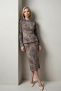 A woman wearing a matching long-sleeve knit top and skirt set with a textured, abstract floral pattern in muted gray, brown, and rust tones, posing in a corner against a light-colored wall.