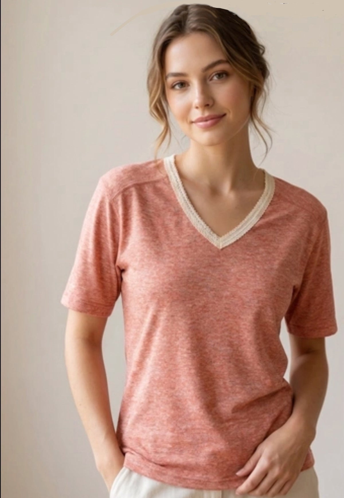 A woman wearing a heathered coral pink short-sleeve V-neck t-shirt with a contrasting cream lace trim, set against a plain beige background.