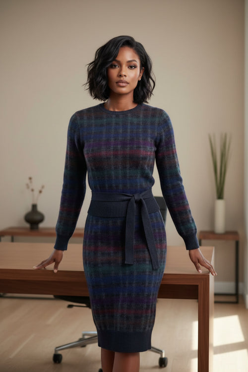 A woman stands in a bright office setting next to a wooden desk, wearing a long-sleeve, knee-length plaid dress in dark blue, gray, and red tones.